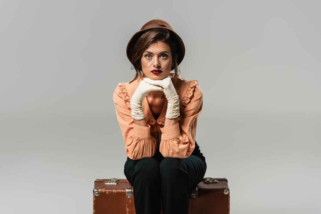 pretty, trendy woman sitting on vintage suitcase while looking at camera on grey - Photo, Image