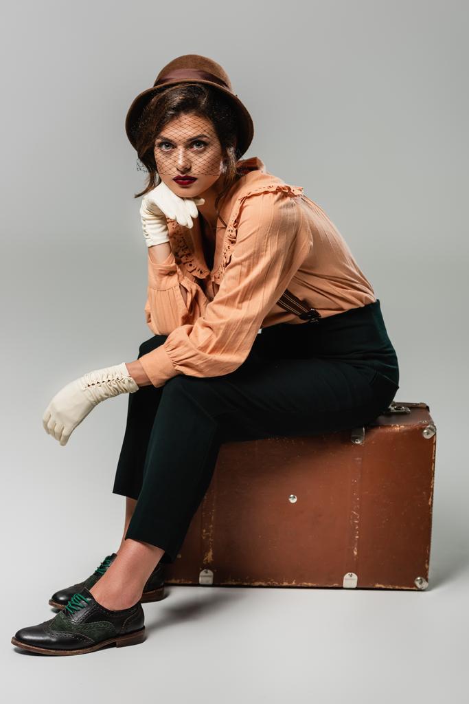 jovem mulher de chapéu sentado na mala vintage com a mão perto do rosto no cinza - Foto, Imagem