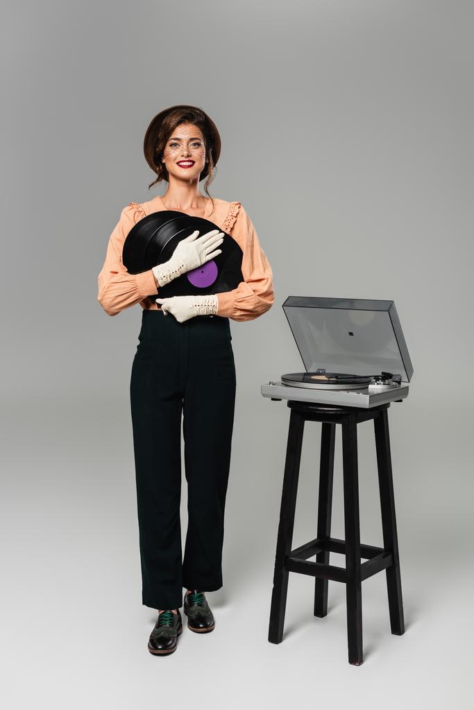mulher elegante feliz em roupas vintage segurando discos de vinil enquanto posando perto gravador no cinza - Foto, Imagem