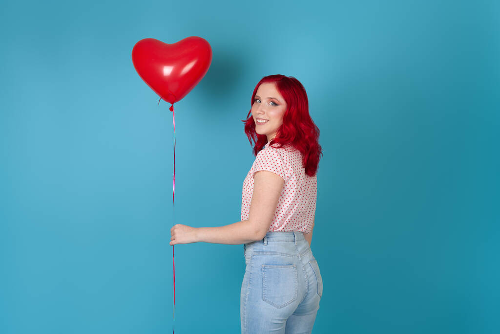 portret zachwyconej pięknej młodej kobiety z rudymi włosami trzymającej czerwony latający balon w kształcie serca, odizolowanej na niebieskim tle - Zdjęcie, obraz