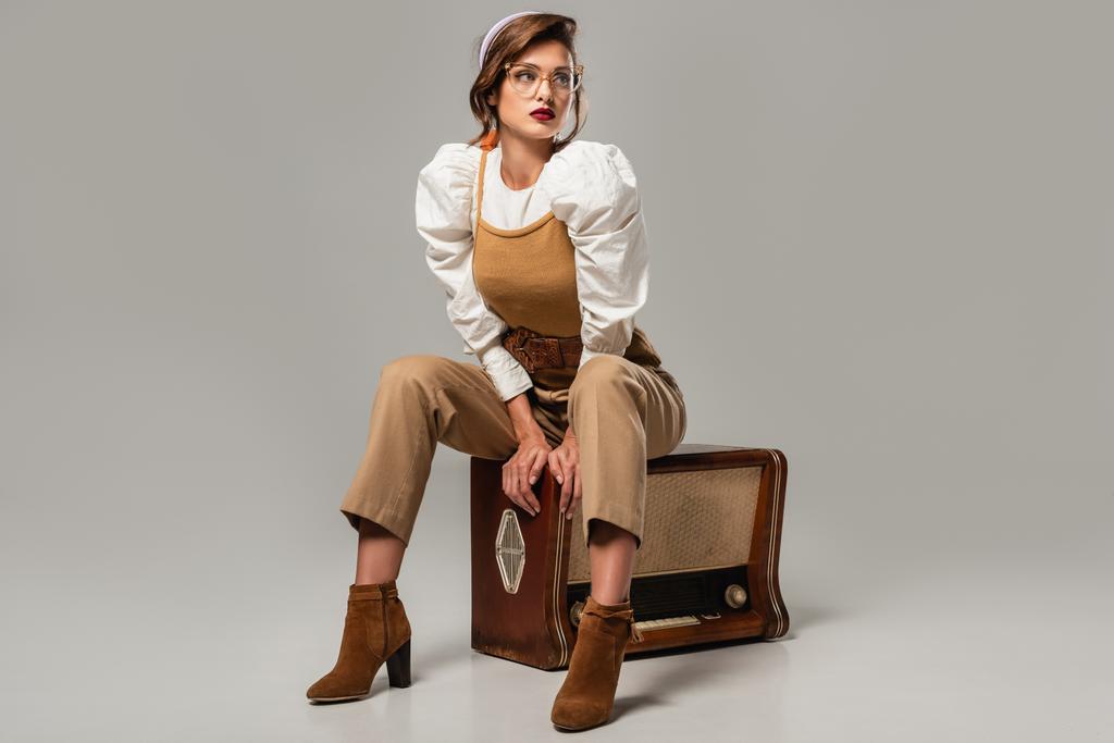 mujer bonita en ropa vintage de moda mirando hacia otro lado mientras está sentado en el receptor de radio vintage en gris - Foto, imagen