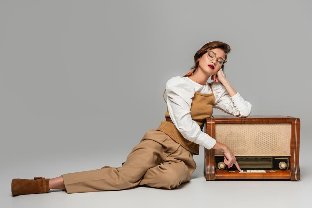 pretty trendy woman pressing button on vintage radio receiver while listening music on grey - Photo, Image