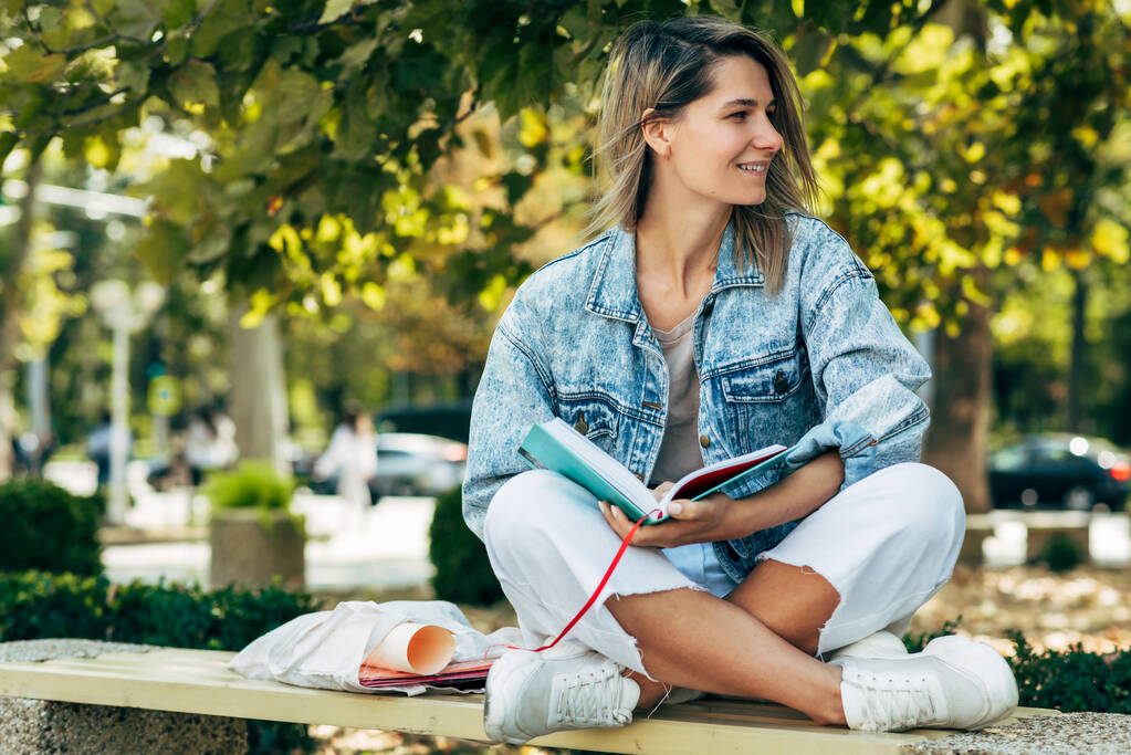 Horizontální obraz šťastné studentky čte knihu, když sedí na lavičce za slunečného dne na městské ulici. Hezká žena v neformálním oblečení si venku odpočine a poučí se z knihy. - Fotografie, Obrázek