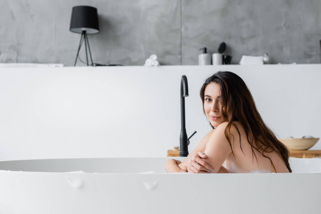 Brunette femme regardant la caméra dans la baignoire à la maison  - Photo, image