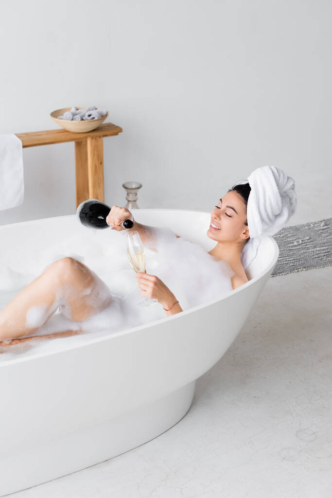 Mulher sorridente derramando champanhe em vidro na banheira  - Foto, Imagem