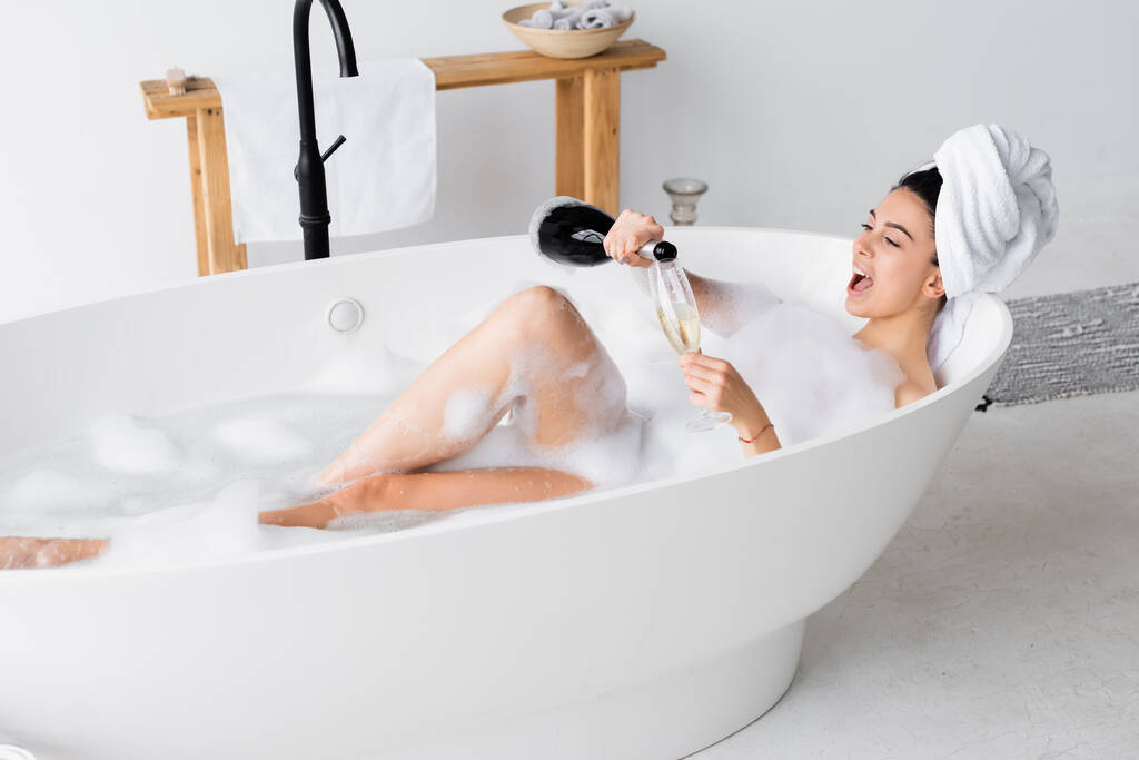 Pozitivní žena nalévání šampaňského ve skle při koupeli  - Fotografie, Obrázek