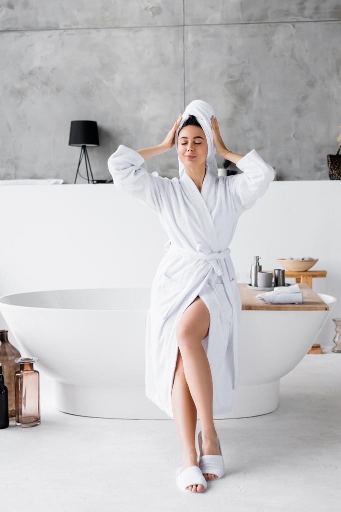 Jonge vrouw in witte badjas en handdoek zitten met gesloten ogen op bad  - Foto, afbeelding