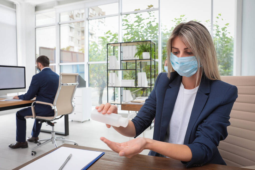 Maskeli bir ofis çalışanı iş yerinde el dezenfektanı kullanıyor. - Fotoğraf, Görsel