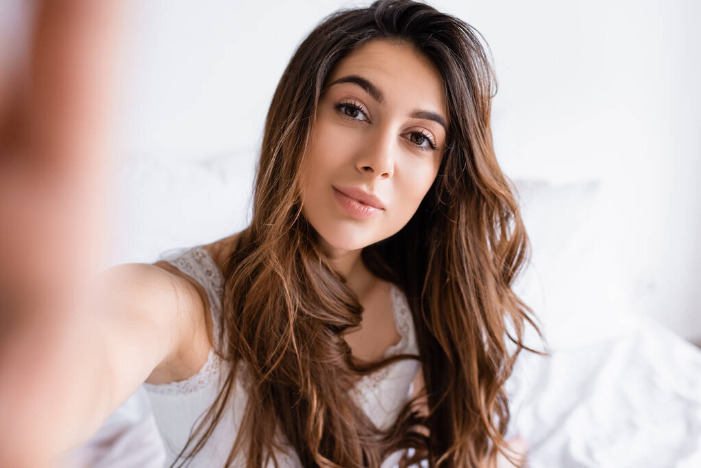 Pijamalı genç bir kadın bulanık arka planda yatak odasında kameraya bakıyor.  - Fotoğraf, Görsel