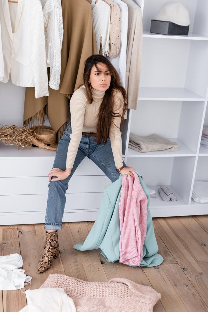 Pensive woman holding clothes while sitting on shelf in wardrobe  - Photo, Image