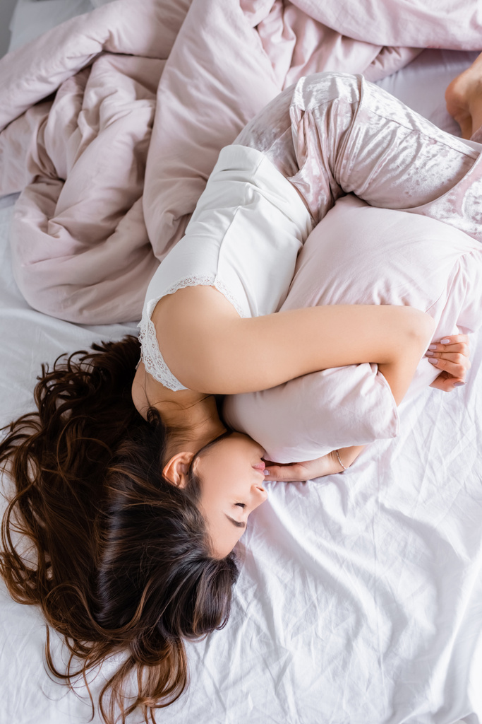 Vista dall'alto della donna bruna in pigiama che abbraccia il cuscino mentre si sdraia sul letto  - Foto, immagini