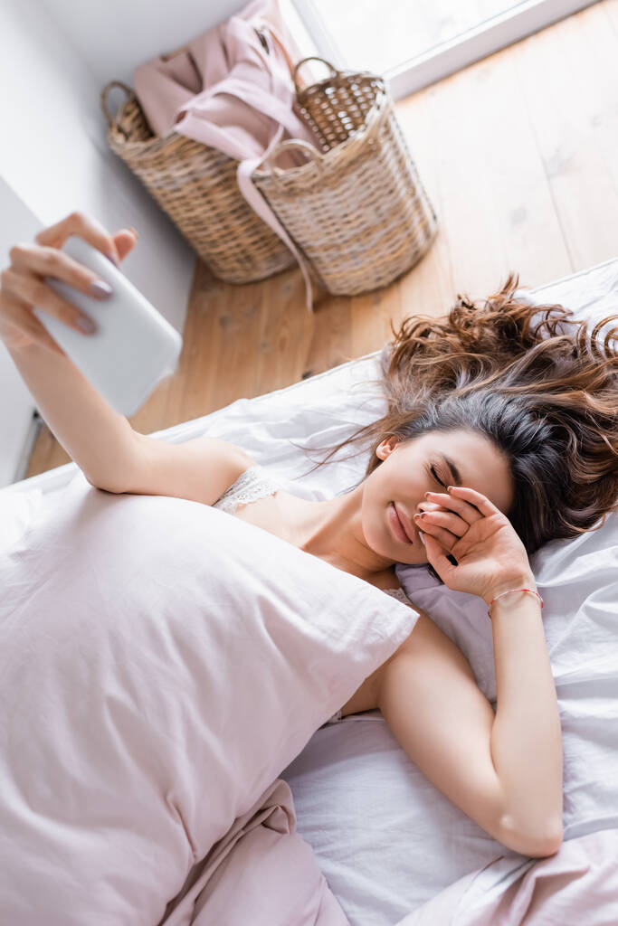 slaperige vrouw het nemen van selfie op smartphone op wazig voorgrond op bed  - Foto, afbeelding