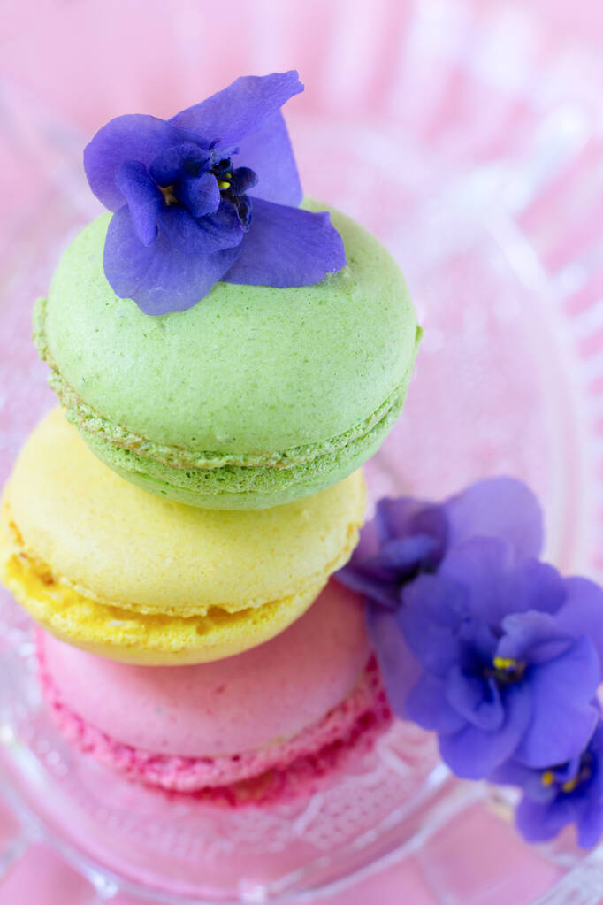 Pile of macaroons decorated with african violet flowers on transparent glass plate.Still life in vertical format with traditional french pastry - Photo, Image