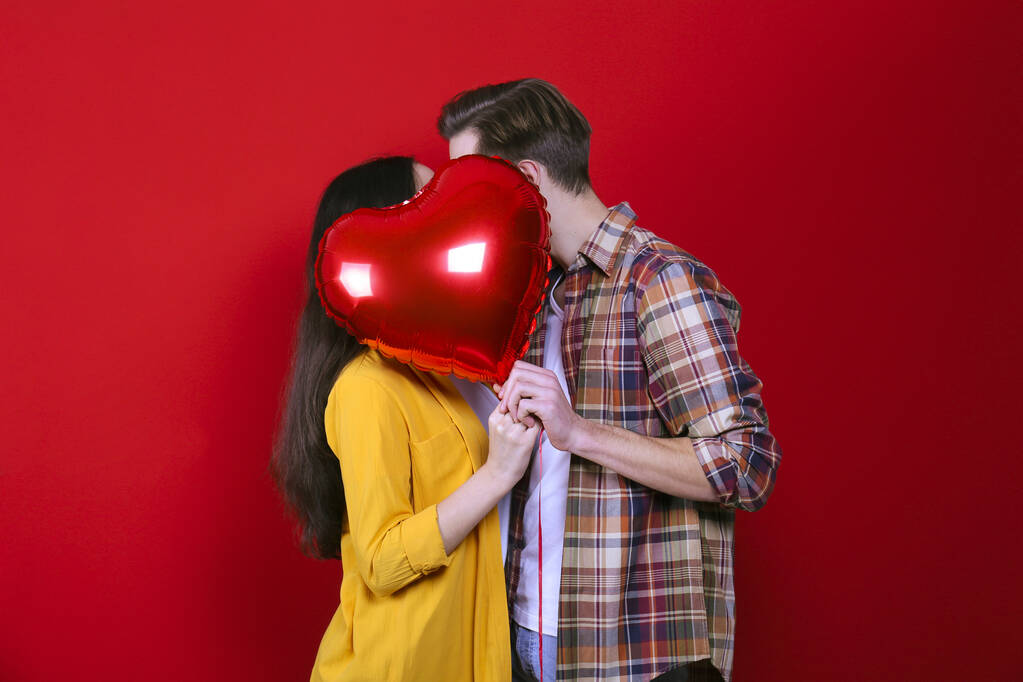 Happy Valentine's day concept. Studio shot of couple in love holding a heart shaped balloon, showing affection. 14th february - the lovers day. Red wall background, copy space, front view portrait. - Photo, Image