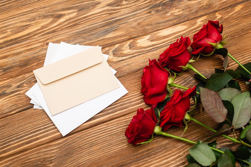 vysoký úhel pohledu na červené růže v blízkosti obálky a papíry na dřevěném povrchu  - Fotografie, Obrázek