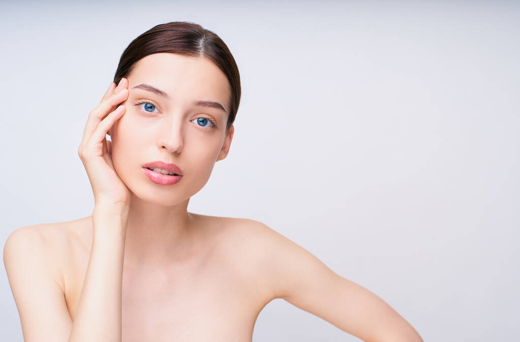 A cute beautiful gentle young girl with beautiful healthy skin, with dark hair and blue eyes, holds her right hand on her face and looks straight. The concept of young and healthy skin, beauty, spa. - Photo, Image