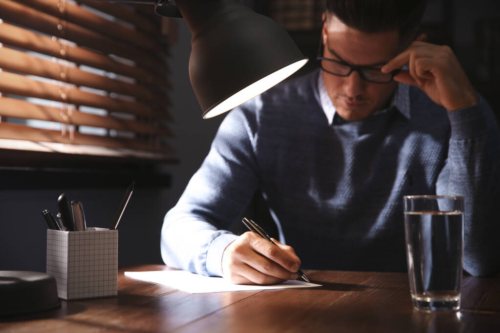 Hombre escribiendo carta en la mesa de madera en habitación oscura, enfoque en la mano - Foto, Imagen
