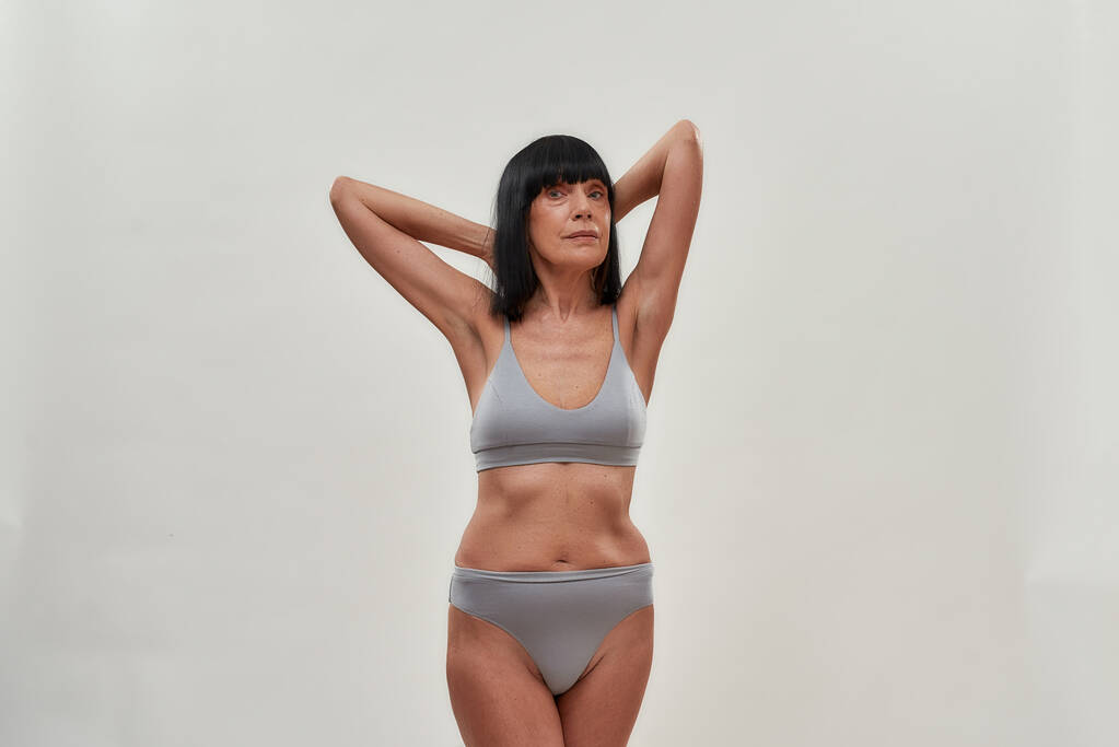 Feeling beautiful. Attractive half naked caucasian senior woman keeping arms raised and looking at camera while posing in lingerie against grey background - Photo, Image