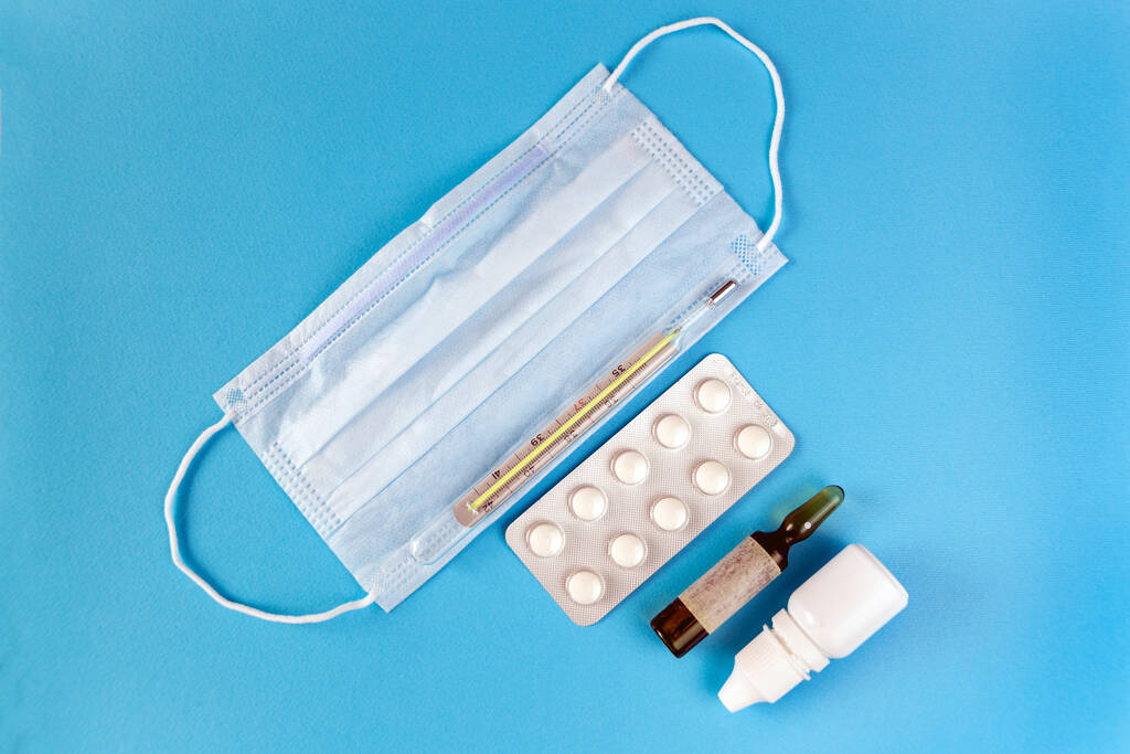 tratamiento médico del virus: mascarilla facial, pastillas, gotas, termómetro sobre el fondo azul - Foto, imagen
