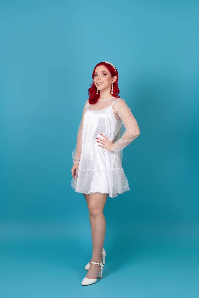 повнометражне усміхнене щасливе красива молода жінка з червоним волоссям у білій сукні з шовкової сітки та білі туфлі на підборах ізольовані на синьому фоні
 - Фото, зображення