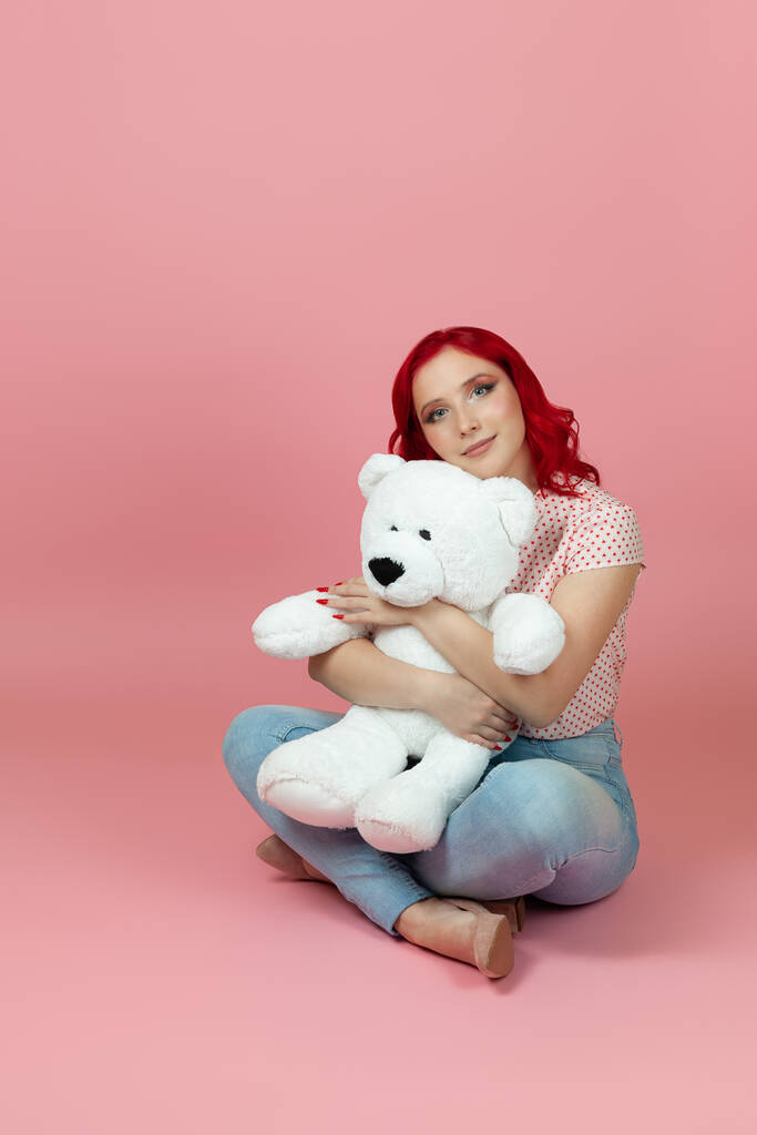 милая, красивая женщина в джинсах с рыжими волосами обнимает большого белого плюшевого медведя, сидящего на полу, изолированного на розовом фоне - Фото, изображение
