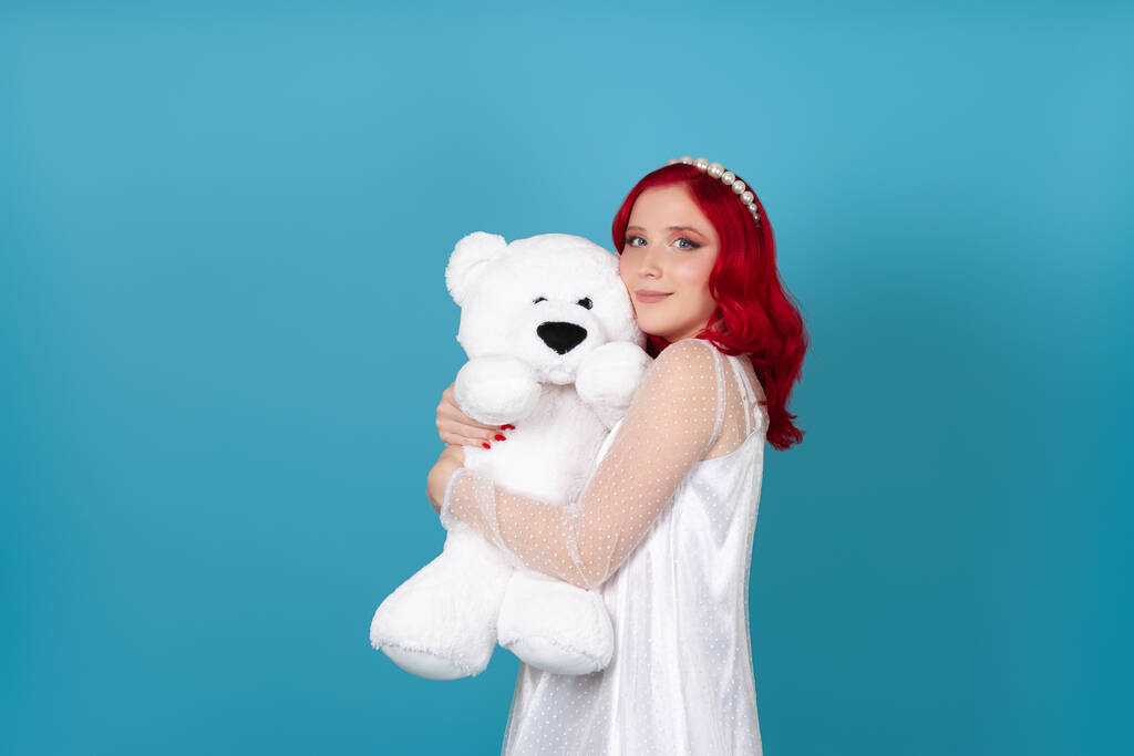 portret in profiel van een mooie jonge vrouw in een witte jurk met rood haar omhelzing van een witte teddybeer geïsoleerd op een blauwe achtergrond - Foto, afbeelding