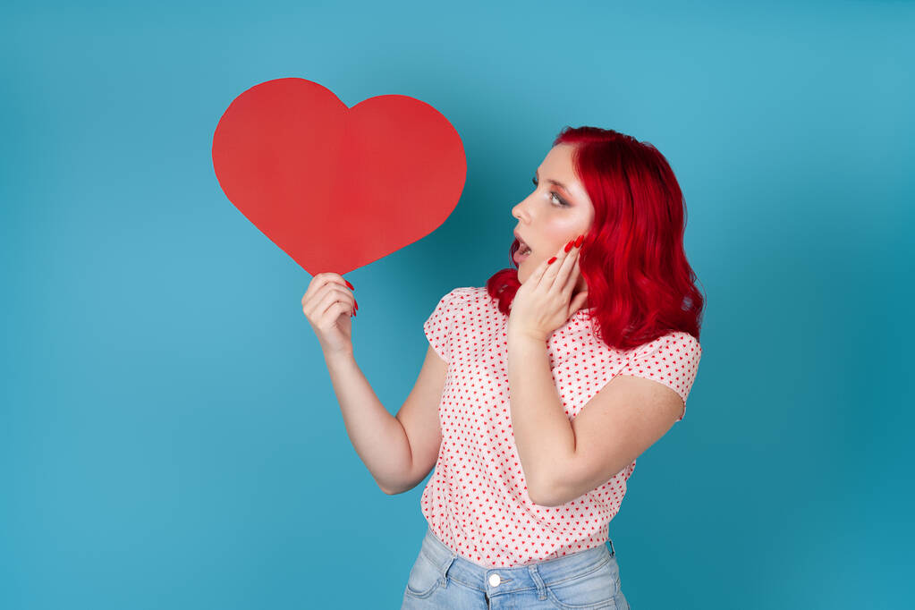 järkyttynyt, innostunut nuori nainen, jolla on punaiset hiukset ja avoin suu, pitää suurta punaista paperia sydäntä eristettynä sinisellä taustalla - Valokuva, kuva