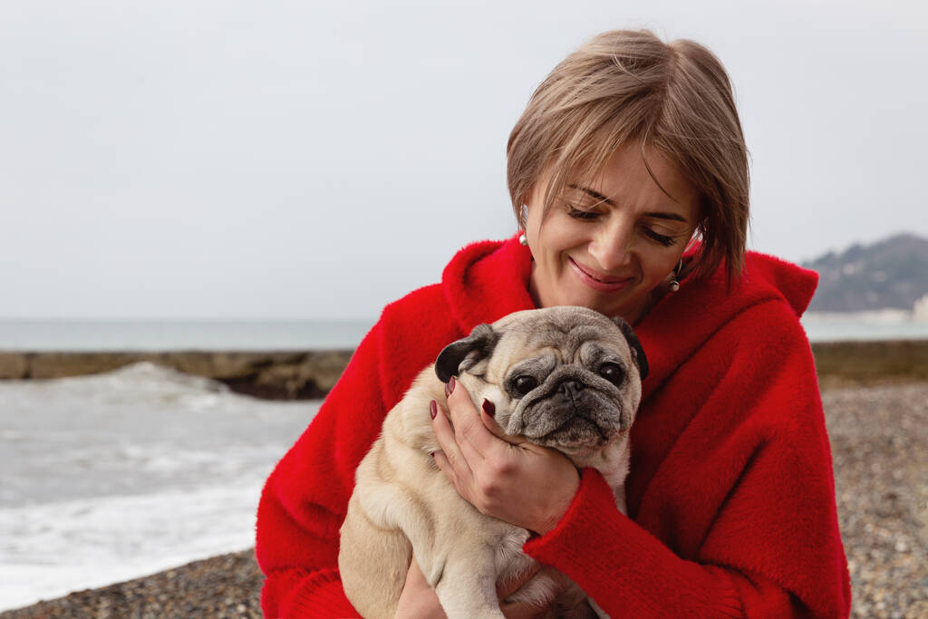A woman with her favorite pug in her arms. Portrait by the sea in winter. - Photo, Image