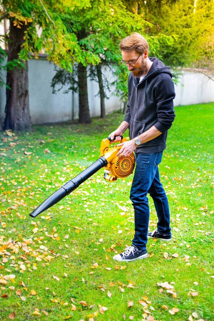 Een jonge man met een baard die een draadloze bladblazer gebruikt in een tuin. Tuinwerkzaamheden. Herfst, herfsttuinieren in een achtertuin, op een gazon, gras. - Foto, afbeelding