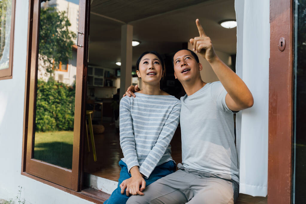 Genç yetişkin Asyalı bir çiftin portresi yukarı bakarken bir adam evin arka planını işaret ediyor. 30 'lu yaşlarda başarılı bir karı koca. Emlak ve emlak başarısı kavramı. - Fotoğraf, Görsel