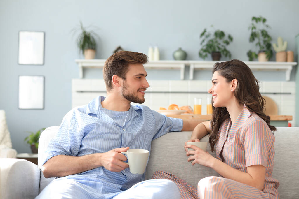 Hermosa pareja joven en pijama se mira y sonríe en un sofá en la sala de estar - Foto, Imagen