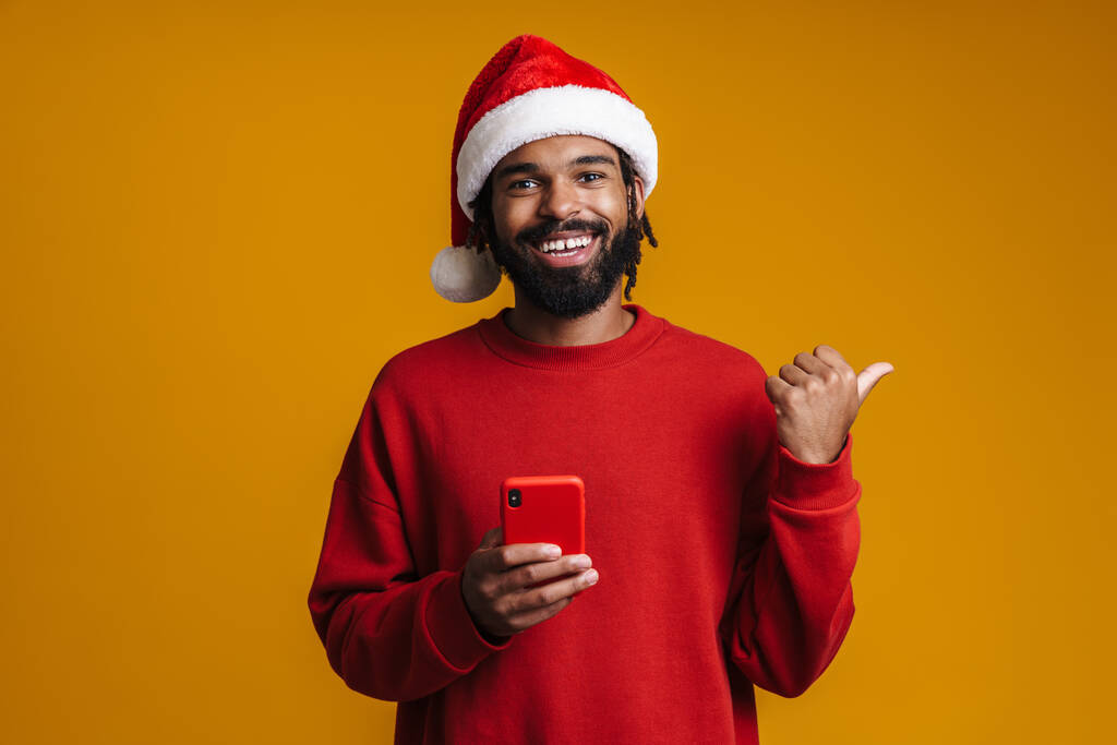 Mutlu Afro-Amerikalı adam sarı arka planda izole edilmiş cep telefonu kullanırken parmağıyla işaret ediyor. - Fotoğraf, Görsel