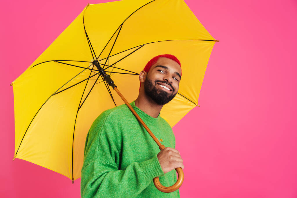 Radosny Afroamerykanin uśmiecha się podczas pozowania z parasolem odizolowanym na różowym tle - Zdjęcie, obraz