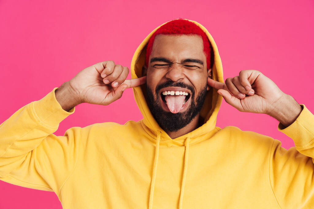 Διασκεδάζοντας με έναν Αφροαμερικανό που δείχνει τη γλώσσα του και βουλώνει τα αυτιά του απομονωμένα σε ροζ φόντο - Φωτογραφία, εικόνα