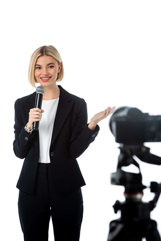 sonriente comentarista rubia con micrófono apuntando con la mano cerca de la cámara digital en primer plano borroso aislado en blanco - Foto, Imagen