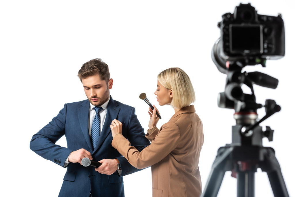 blonde makeup artist with cosmetic brush near news anchor buttoning his blazer isolated on white, blurred foreground - Photo, Image