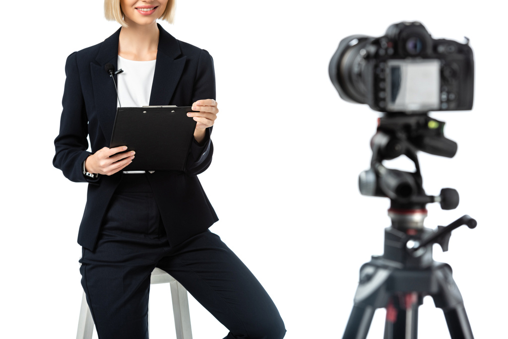 oříznutý pohled na usmívající se moderátorku se schránkou sedící poblíž digitálního fotoaparátu na rozmazaném popředí izolovaném na bílém - Fotografie, Obrázek