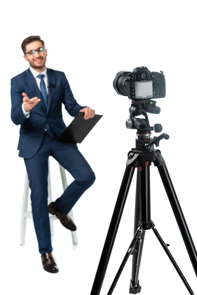 fröhlicher Nachrichtenanker, der mit der Hand zeigt, während er auf einem hohen Hocker neben der Digitalkamera auf weißem, verschwommenem Vordergrund sitzt - Foto, Bild