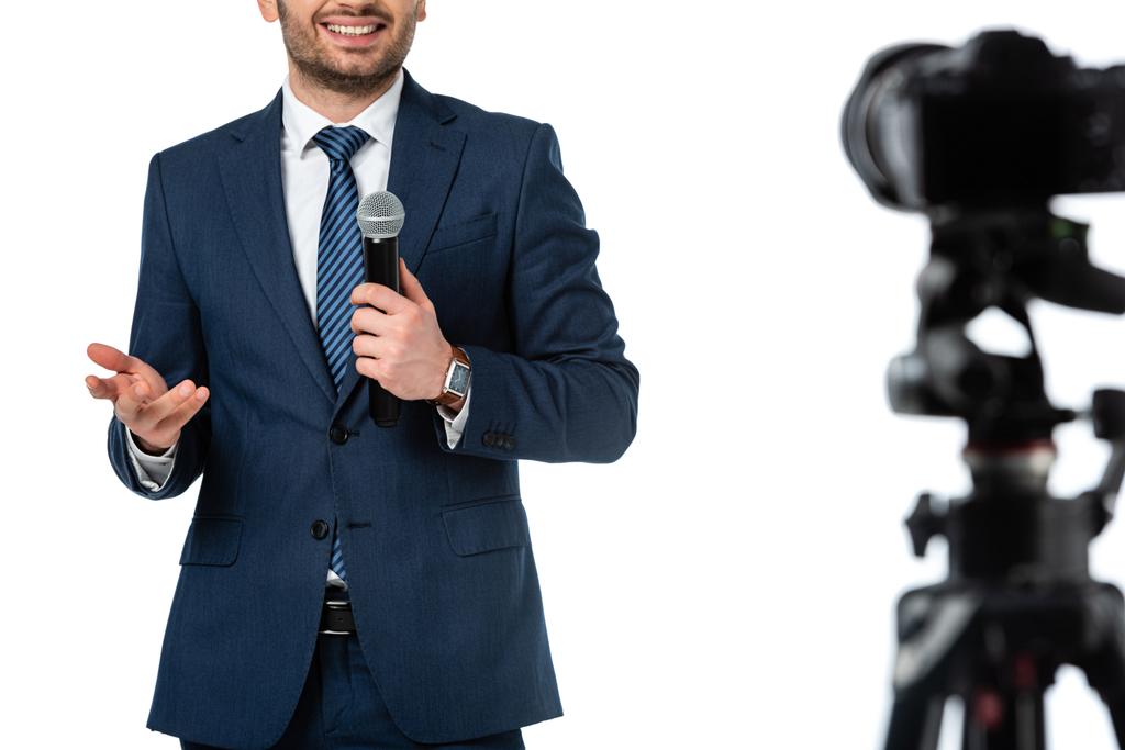 oříznutý pohled na usmívající se zpravodajskou kotvu s mikrofonem v blízkosti digitálního fotoaparátu izolovaného na bílém rozmazaném popředí - Fotografie, Obrázek