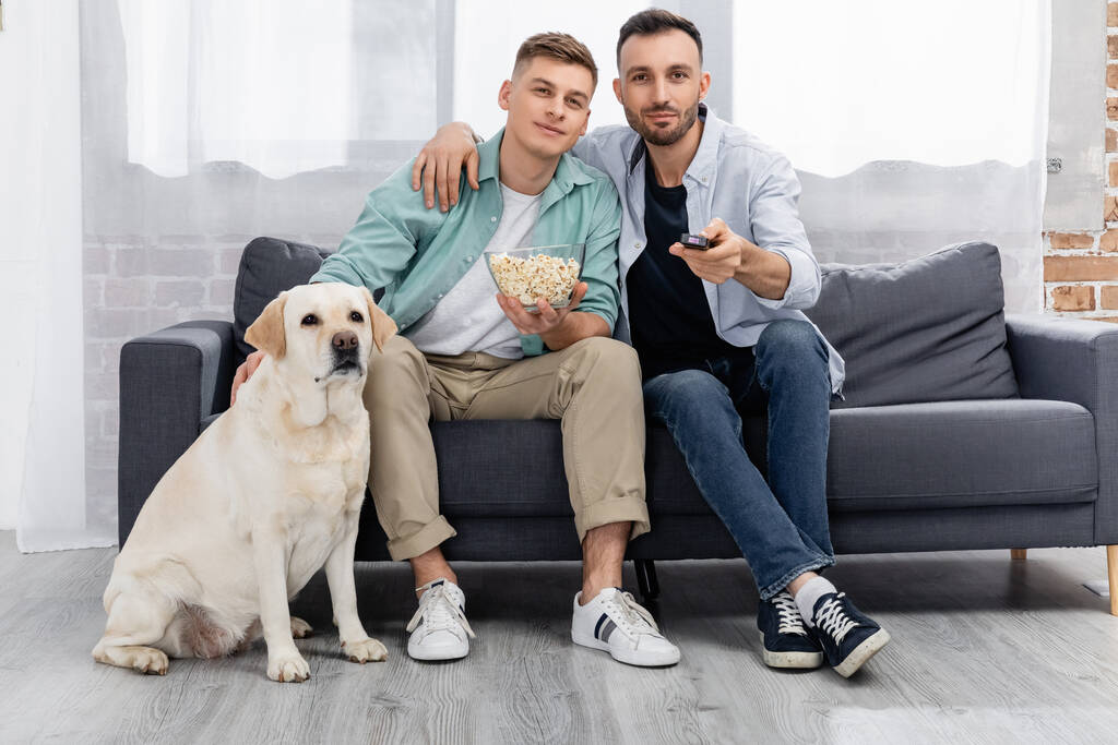 fröhliches gleichgeschlechtliches Paar schaut Film mit Popcorn bei Labrador  - Foto, Bild