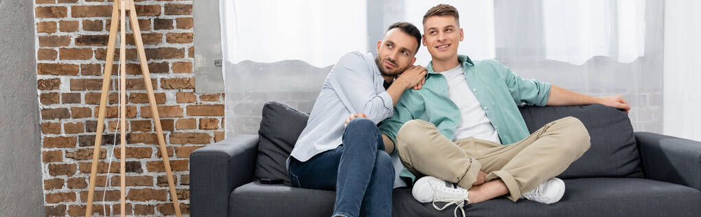 gai même sexe couple regardant loin tandis que assis sur canapé à la maison, bannière - Photo, image