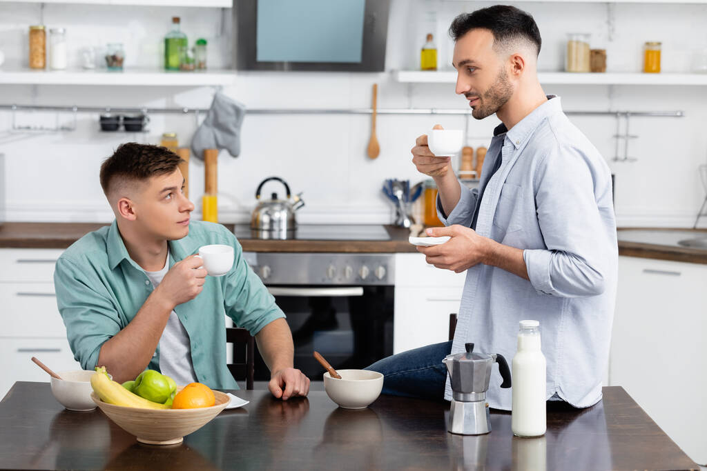 feliz pareja del mismo sexo mirándose mientras sostiene tazas en la cocina  - Foto, imagen