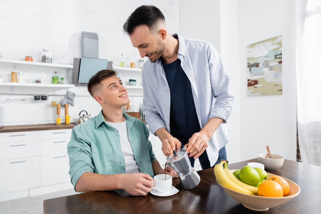happy homosexual man pouring coffee near husband in kitchen - Photo, Image