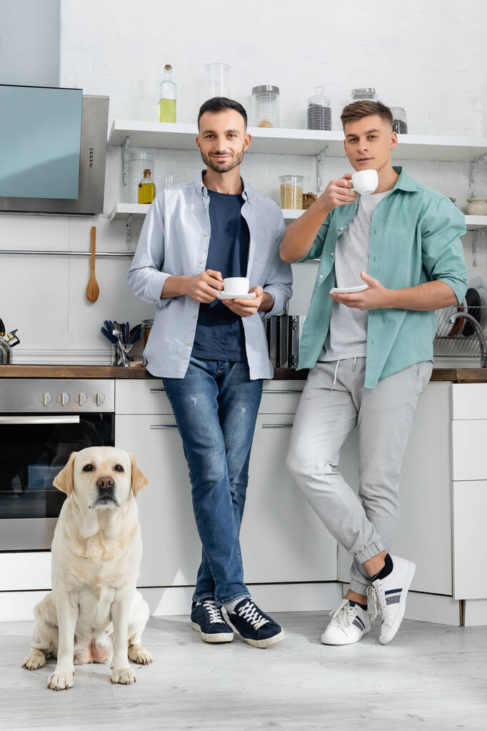 幸せな同性愛者の男性がカップを持ってキッチンで犬の近くに立っています  - 写真・画像