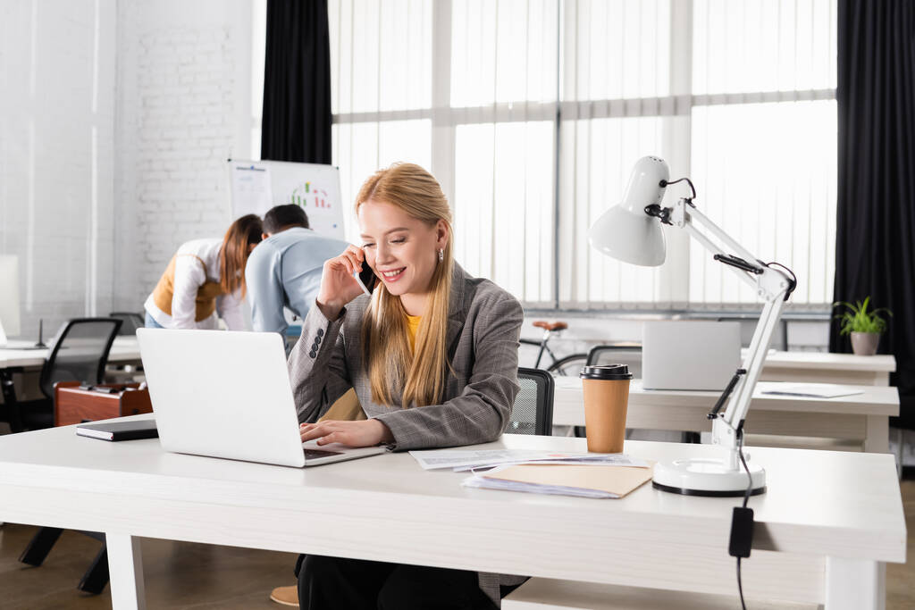 Улыбающаяся деловая женщина разговаривает на смартфоне возле ноутбука и кофе, чтобы пойти в офис  - Фото, изображение