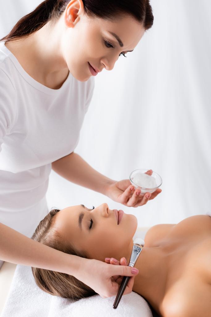 esteticista segurando escova cosmética e recipiente com máscara facial perto da mulher com os olhos fechados deitados na mesa de massagem - Foto, Imagem