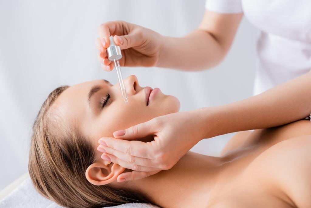 Kosmetikerin hält Pipette und appliziert Serum auf das Gesicht einer Frau mit geschlossenen Augen im Wellness-Salon  - Foto, Bild