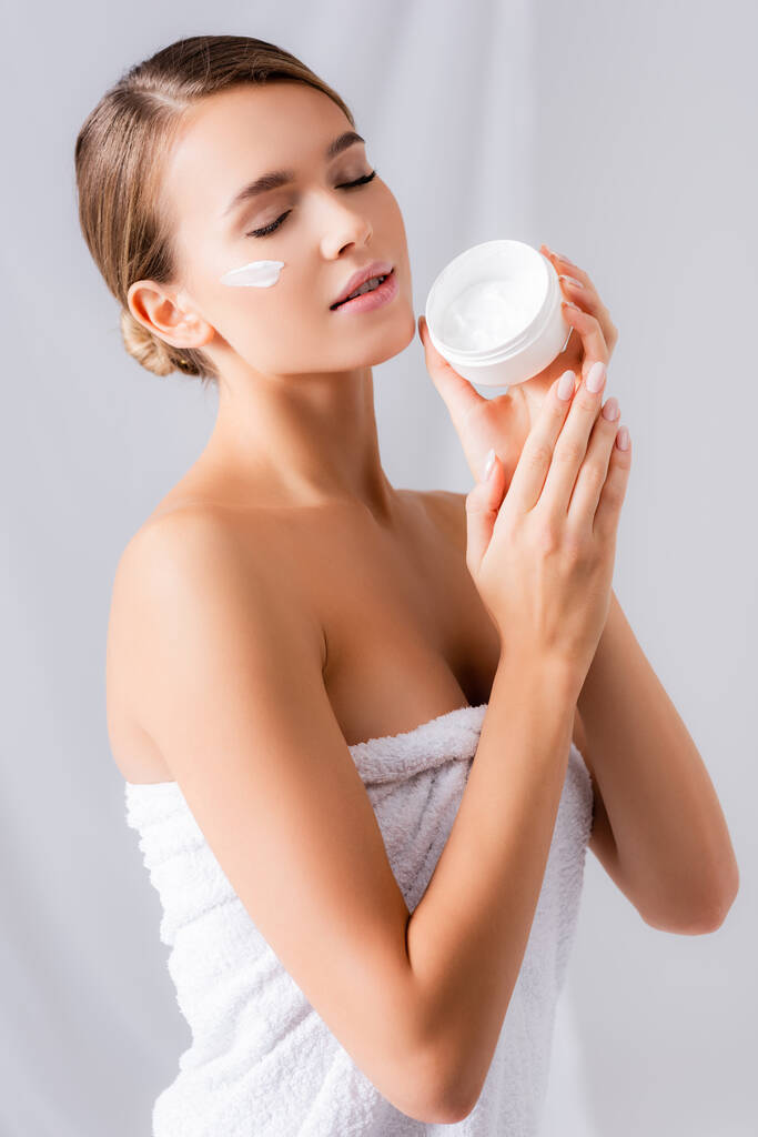 jeune femme avec les yeux fermés et crème sur le visage tenant pot sur blanc - Photo, image