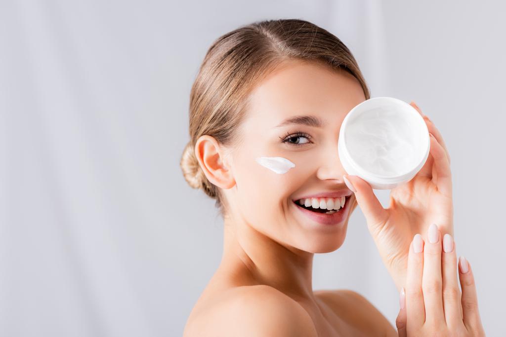 joyeuse jeune femme avec de la crème sur le visage tenant le pot et regardant la caméra sur blanc - Photo, image