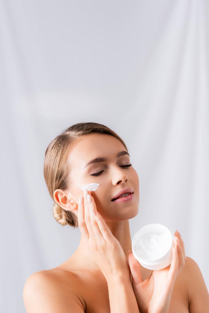giovane donna con spalle nude che tiene il vaso e applica la crema per il viso sul bianco - Foto, immagini